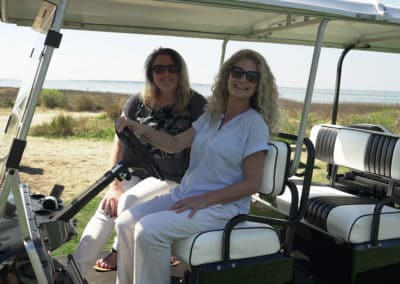 Two women in sunglasses sit together in a golf cart, smiling and enjoying their time outdoors in a low-speed vehicle.